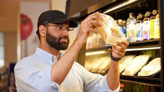 Person examining a packaged food item in front of a refrigerated display with beverages and snacks.
